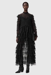 Svart spetsklänning med lager av volanger, långa genomskinliga ärmar och hög halsringning. Stylad med knähöga svarta stövlar och en liten kuvertväska.