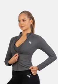 Šedá športová bundy na zips s dlhými rukávmi, úzky strih, rebrovaná textúra a biele logo na ľavom ramene. Nosená cez zhodný top.