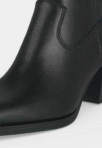 Gros plan d'une botte cheville en cuir noir avec un bout pointu, des coutures visibles et un talon épais sur un fond clair.