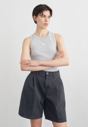 Ung person med kort mørkt hår iført en grå ærmeløs top og løse sorte shorts, armene krydsede, kigger til siden.