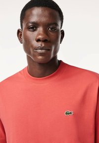 T-shirt a maniche corte di colore corallo, realizzata in cotone, con un colletto a costine. Presenta un logo di coccodrillo ricamato di colore verde sul lato sinistro del petto.