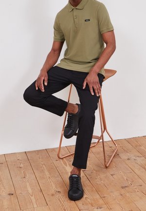 Man zit op een koperen kruk en draagt een olijfgroen poloshirt, zwarte broek en zwarte Calvin Klein sneakers op een houten vloer.