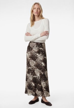 Kobieta stojąca z założonymi ramionami, ubrana w biały top z długim rękawem, długą brązowo-kremową spódnicę w wzory oraz ciemne baleriny.