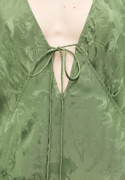 Πράσινο φόρεμα με βαθύ V λαιμόκοψη, λεπτομέρεια κορδονιού στο κέντρο και υφασμάτινη υφή με ανάγλυφο σχέδιο αλόγου σε όλο το μήκος.