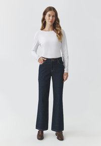 Jeans acampanados de color azul oscuro con una cintura alta, cinco bolsillos y costuras de contraste, combinados con una sencilla blusa blanca de mangas largas.