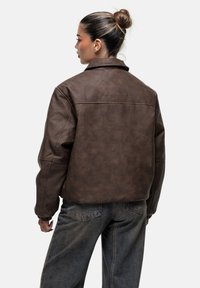 Giacca in finta pelle marrone con una vestibilità rilassata, cuciture visibili e colletto. Presenta una finitura texturizzata e un logo discreto sul retro.