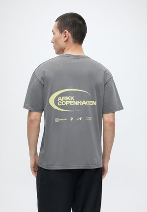 Άνδρας με γυρισμένη πλάτη, φοράει γκρι μπλουζάκι ARKK Copenhagen με μεγάλο κίτρινο λογότυπο και εικονίδια στην πλάτη, συνδυασμένο με μαύρο παντελόνι.