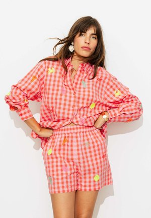 Femme aux cheveux bruns porte une chemise et un short à carreaux corail et blancs assortis avec des motifs brodés colorés, debout les mains sur les hanches.