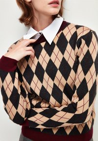 Maglione argyle multicolore con beige, nero e bordeaux. Presenta un colletto e polsini a coste bordeaux. Texture morbida con motivi a rombi.