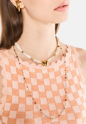Gold heart pendant necklace on textured beige and orange checkered top, with multi-colored gemstones strung on a delicate gold chain.