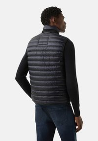 Czarna kamizelka puchowa z poziomym pikowaniem, wysokim kołnierzem i błyszczącym wykończeniem. Noszona na czarnej bluzce z długim rękawem, zestawiona z niebieskimi dżinsami.