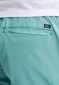 Pantalons en coton turquoise avec une poche arrière ornée d'une couture et d'un petit étiquette de marque. Texture lisse et design décontracté.
