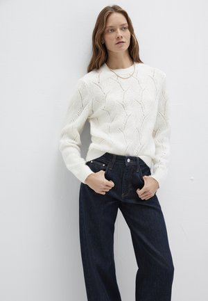 Weißer Strickpullover mit einem strukturierten Wellenmuster, kombiniert mit hoch taillierter dunkler Denim-Jeans. Das Modell steht mit den Händen in den Taschen.