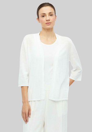 Frau mit braunen, nach hinten gebundenen Haaren, die eine weiße, offene Strickjacke, ein weißes Top und weiße Hosen trägt und vor einem grauen Hintergrund steht.