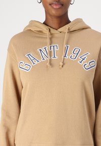 Haut à capuche beige avec cordon de serrage, présentant un design à l'avant avec l'inscription "GANT 1949" en broderie bleue. Texture de tissu douce, coupe décontractée.
