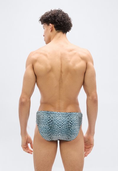 Άνδρας με σγουρά σκούρα μαλλιά, στέκεται με την πλάτη στην κάμερα, φοράει μπλε και μαύρο μαγιό με λεοπάρ σχέδιο, σε απλό λευκό φόντο.