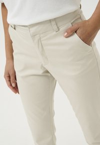 Beżowe spodnie z gładkiego materiału, z zamkiem z przodu, bocznymi kieszeniami i dopasowanym krojem widocznym w okolicy pasa i bioder.