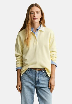 Jonge vrouw met lang blond haar, gekleed in een lichtgele trui over een blauw overhemd met kraag en lichtblauwe spijkerbroek met een bruine riem.