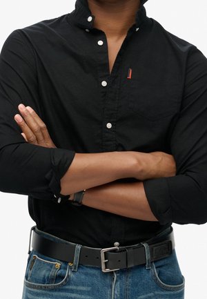 Mand med armene krydset iført en sort skjorte med knapper, opkørte ærmer, sort læderbælte og blå jeans mod en hvid baggrund.