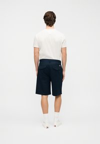 Σκούρες μπλε σορτς από βαμβακερό μείγμα, με επίπεδη μπροστινή όψη, δύο πίσω τσέπες με καπάκια και κλασική εφαρμογή. Φοριούνται με λευκή μπλούζα.