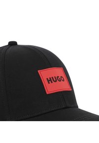 Μαύρο καπέλο με δομημένο σχήμα, λείο υλικό και κόκκινη ελαστική ετικέτα με ανάγλυφο το λογότυπο "HUGO" στο μπροστινό μέρος.