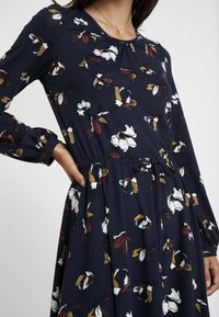 Robe florale à manches longues, fond marine, avec des fleurs blanches et bordeaux, encolure froncée, taille élastique et détail à cordon.
