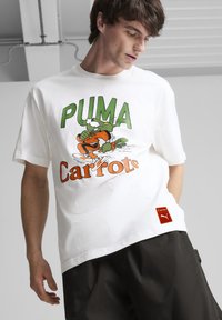 Balta kokvilnas t-krekls ar zaļu un oranžu multfilmu grafiku, kurā virsū rakstīts "PUMA" un apakšā "Carrots", un sarkanā etiķete apakšmalā.