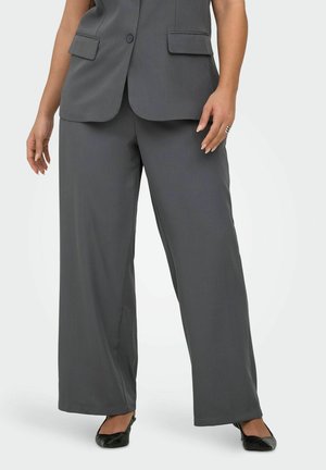 Kvinde iført mørkegrå bukser med vide ben, matchende knapforet blazer, sorte flade sko og en sølvring på højre hånd.