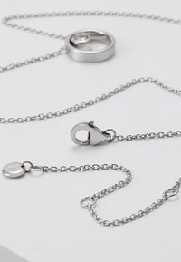 Collier en chaîne argentée avec un pendentif rond orné d'une pierre transparente, et un fermoir mousqueton. Texture lisse avec un design en chaîne maillée.