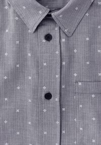 Camisa de patrón en espiga gris con pequeños acentos blancos. Cuenta con un cuello clásico, botones negros y un bolsillo en el pecho.