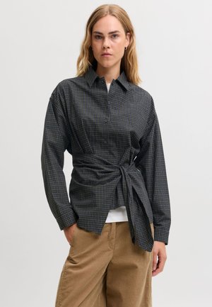 Un mannequin aux cheveux blonds jusqu’aux épaules porte une chemise à carreaux foncés nouée à la taille par-dessus un haut blanc et un pantalon beige, sur un fond uni.