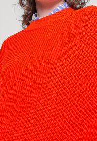 Sweter dziany w jasnym pomarańczowym kolorze, z teksturową powierzchnią, szerokim okrągłym dekoltem i ściągaczami. Jest noszony na wzorzystej koszuli.