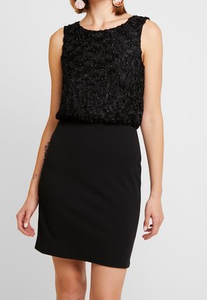 Ärmelloses schwarzes Kleid mit strukturiertem, perlenbesetztem Oberteil und glattem, tailliertem Rock, getragen von einer Person mit kurzen Haaren und großen runden Ohrringen.