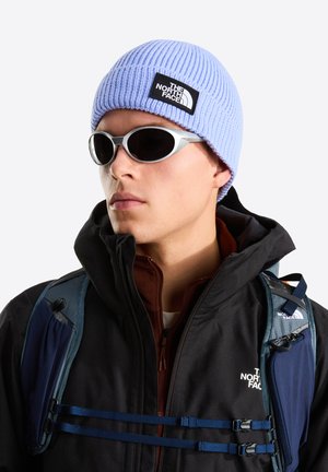 Lyseblå ribstrikket hue med logo-patch, hvid lynlåstrøje, brun indvendig lag, grå sportslige solbriller og blå rygsæksremme.