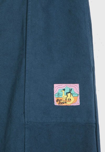 Tissu bleu avec un petit écusson carré montrant un chameau portant un chapeau et portant une échelle, avec un arrière-plan et un texte stylisés.