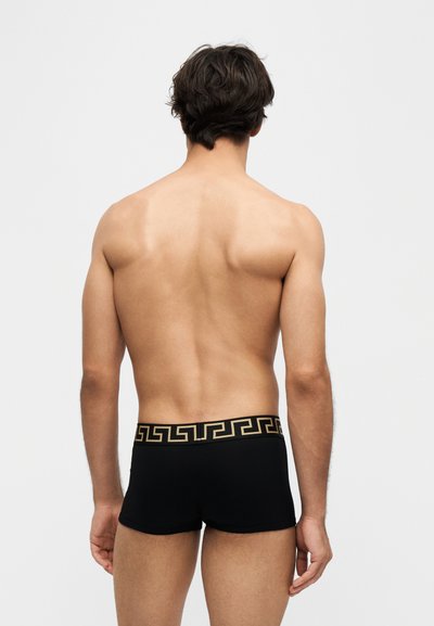 Homme torse nu aux cheveux foncés, debout de dos, portant un boxer noir avec une ceinture dorée à motif sur un fond uni.