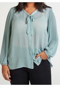 Blusa di un delicato verde chiaro realizzata in tessuto trasparente con piccoli motivi a pois, scollo a V, chiusura con bottoni davanti e dettaglio con fiocco al collo, maniche lunghe plissettate.