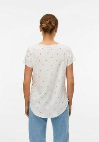 Hvid bluse med små røde mønstre, korte ærmer, afrundet kant og glat tekstur, båret med lyseblå jeans.