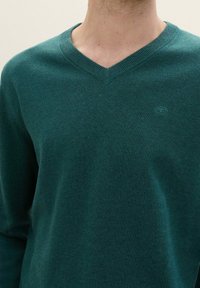 Pulover cu guler în V din material tricotat de culoare teal închis. Are guler striat și un accent logo mic pe piept. Textură moale.