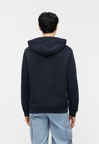 Άνδρας που στέκεται με την πλάτη γυρισμένη, φορώντας μπλε τούρκου ζακέτα με κουκούλα και ανοιχτόχρωμο γαλάζιο παντελόνι με πλαϊνές τσέπες, μπροστά σε απλό λευκό φόντο.