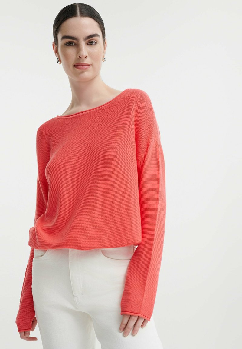 Korallenfarbener gerippter Pullover mit weitem Bootsausschnitt und langen Ärmeln, getragen über weißen hochtaillierten Jeans, mit entspannter Passform.