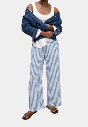 Person iført blå og hvide stribede bukser med vide ben, hvid tanktop, denimjakke uden på skuldrene og brune sandaler mod ensfarvet baggrund.