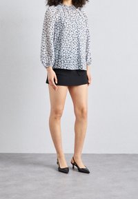 Blouse blanche transparente à imprimé floral, manches ballon ; associée à une mini-jupe noire et des escarpins noirs pointus. Tissu lisse, coupe décontractée.