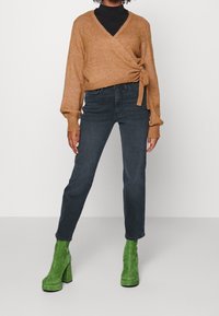 Maglione avvolgente marrone lucido con un nodo, abbinato a jeans di denim scuro e stivali con tacco in camoscio verde, in piedi davanti a uno sfondo neutro.