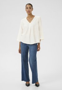 Blouse blanche à col en V avec des manches larges, associée à un jean large bleu. Texture lisse et coupe décontractée. Des chaussures noires à bout pointu complètent la tenue.