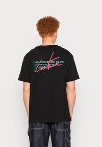 Camiseta de algodón negra con un diseño colorido en la parte posterior, con texto y una firma estilizada en rosa y verde. Diseño casual, de manga corta.