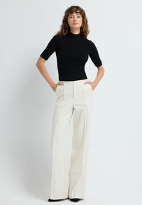 Czarny, prążkowany top z krótkim rękawem w połączeniu z jasnobeżowymi, szerokimi spodniami w kanty o wysokim stanie. Modelka stoi z rękami w kieszeniach.