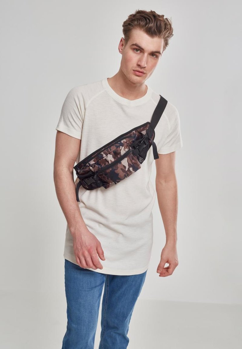 Borsa a fascia mimetica realizzata in tessuto testurizzato, con chiusura a zip e tracolla regolabile, indossata su una maglietta chiara e jeans blu.