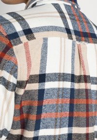 Flanelová košile s velkým klasickým károvaným vzorem v námořnické modři, červené a béžové na bílém pozadí s měkkým, texturovaným povrchem.