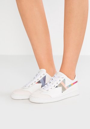 Baskets blanches basses avec des motifs géométriques colorés sur les côtés, portées pieds nus, debout sur un sol blanc uni.